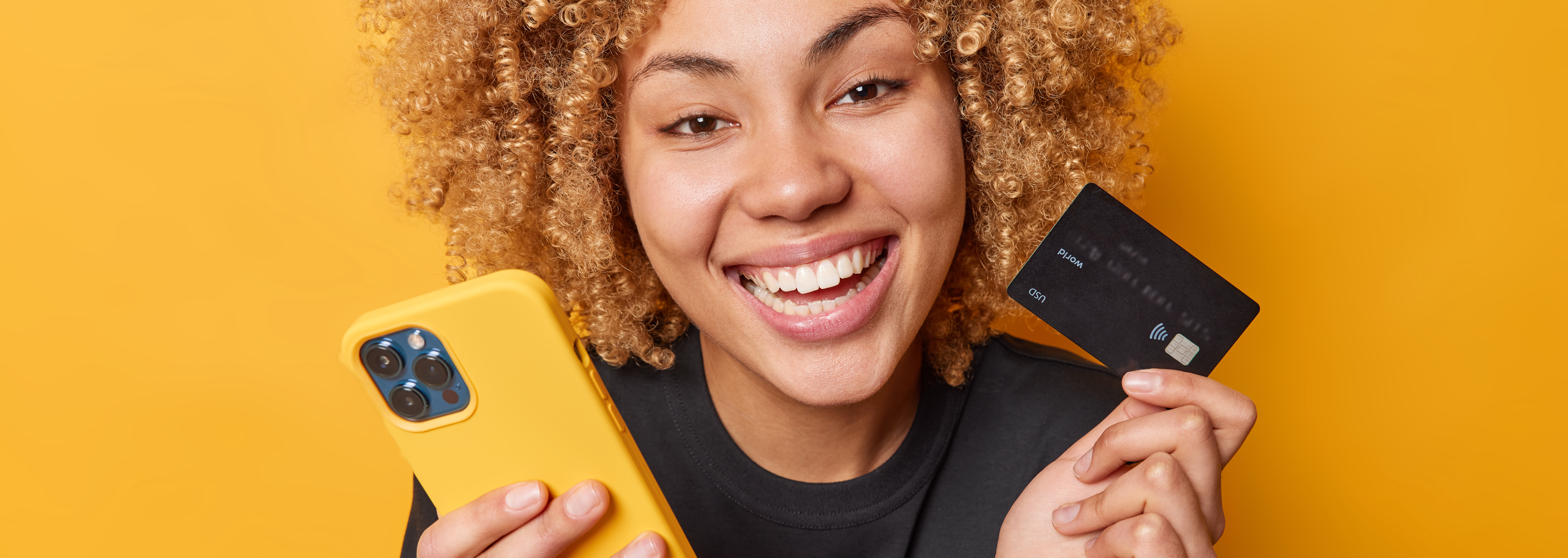 happy-curly-female-shopper-enjoys-easy-paying-goods-online-smiles-broadly-shows-white-teeth-uses-mobile-phone-credit-card-dressed-casual-black-t-shirt-isolated-yellow-background